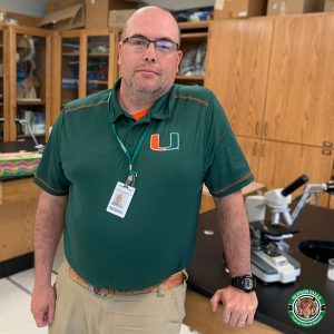 Mr. Delancy, a white man with a clean-shaven head and glasses, stands in a classroom. He wears a green polo shirt and a lanyard. 